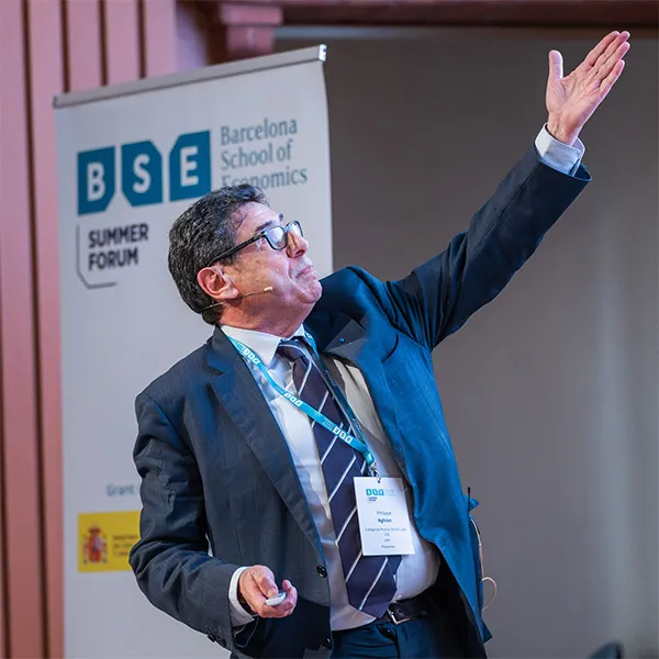 Philippe Aghion stands gestures to a presentation slide. In the background the logo of the BSE Summer Forum and the Spanish Government are partially visible