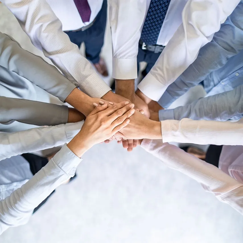 Overhead photo of a group of diverse professionals standing in a circle with their hands stacked together in the center, symbolizing teamwork, unity, and collaboration. The people are wearing business attire, including white, blue, and pink dress shirts, and ties.