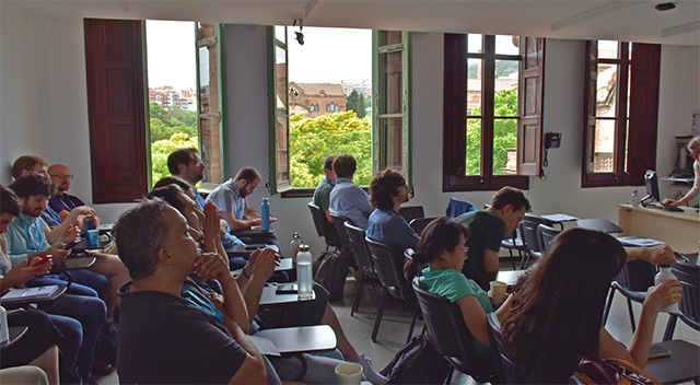 People sit in a classroom. Through the windows, the view includes the bright blue sky and palm trees.