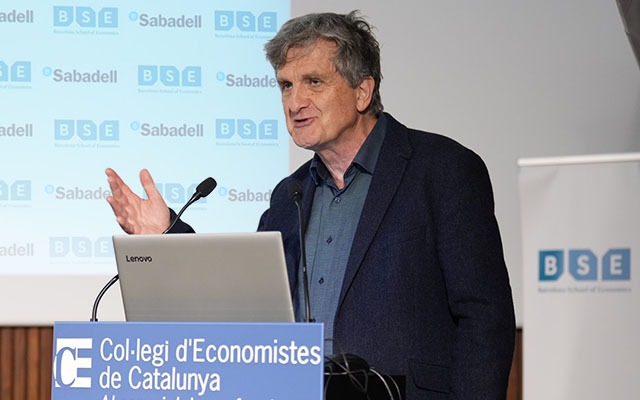Professor John Moore speaks from behind a lectern that displays the logo of the host venue, the Col·legi d'Economistes de Catalunya. Behind him are screens showing the logos of the Barcelona School of Economics and Banc Sabadell.