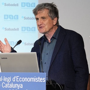 Professor John Moore speaks from behind a lectern that displays the logo of the host venue, the Col·legi d'Economistes de Catalunya. Behind him are screens showing the logos of the Barcelona School of Economics and Banc Sabadell.