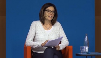 Ada Ferrer-i-Carbonell sits in a red chair in front of a blue backdrop. She is holding notes and speaking. On a table next to her there is a bottle of water and a water glass.