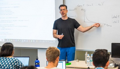 Professor Hannes Mueller gestures to notes written on a whiteboard at the front of a classroom while students watch from their seats