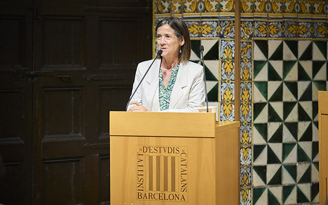 Teresa Garcia-Milà speaks at a wooden lectern decorated with the logo of the Institut d'Estudis Catalans